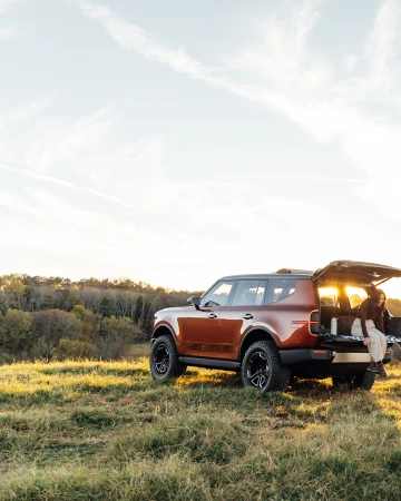 Brown SUV parked in a grassy field at sunset with a person sitting in the open trunk, enjoying the view of the landscape.