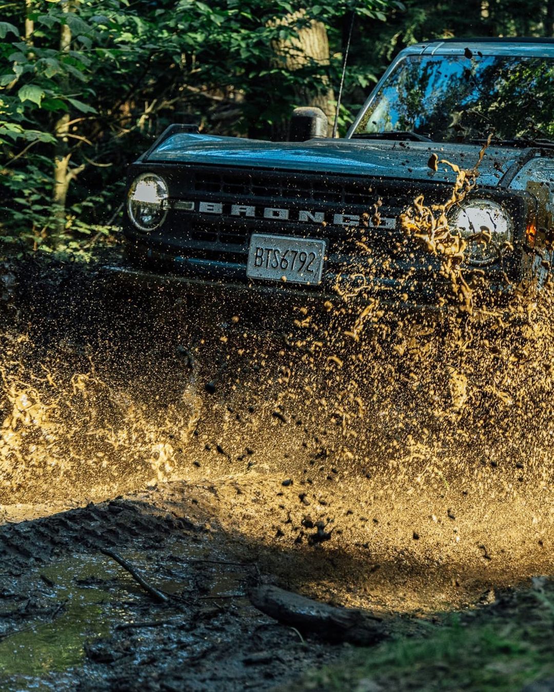 Off-road vehicle splashes through mud on a wooded trail. Dark green Ford Bronco with license plate partially visible, trees in background.