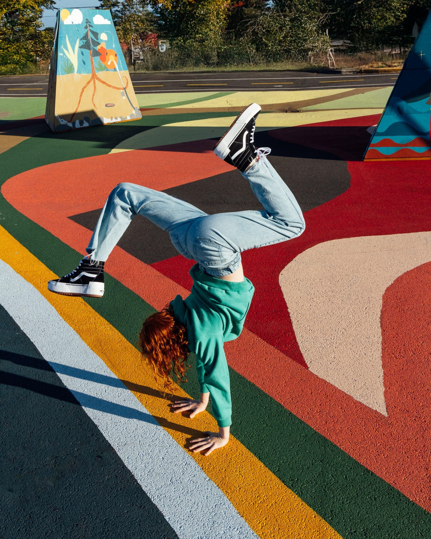 A person in a green sweatshirt and light blue jeans does a handstand wearing platform Vans sneakers on a multicolored background.