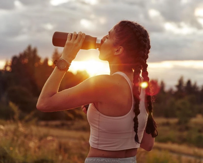 Woman drinking from a water bottle outdoors against a sunset background, wearing a sports bra, wrist watch, and having braided hair.