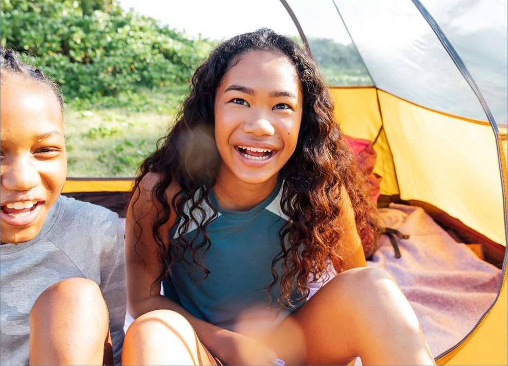 Two smiling children inside a yellow and white tent with greenery in the background, bathed in natural light, conveying a sense of joy.