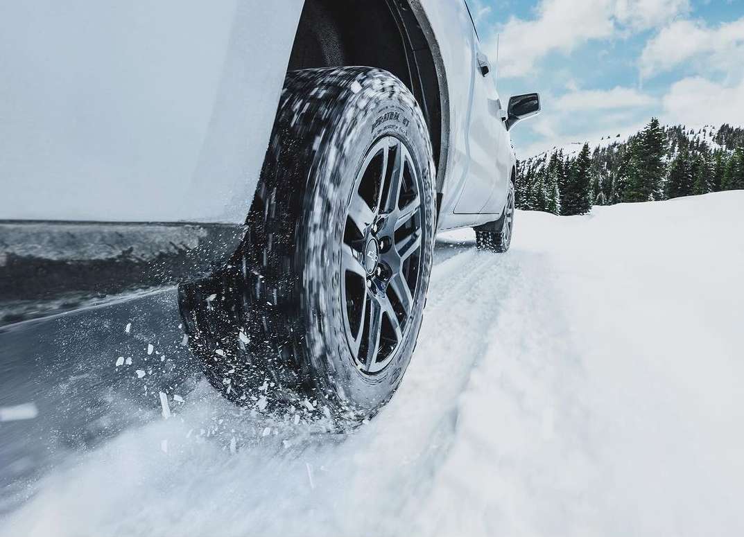 Vehicle tire driving on a snowy road with pine trees in the background under a bright blue sky with some clouds.