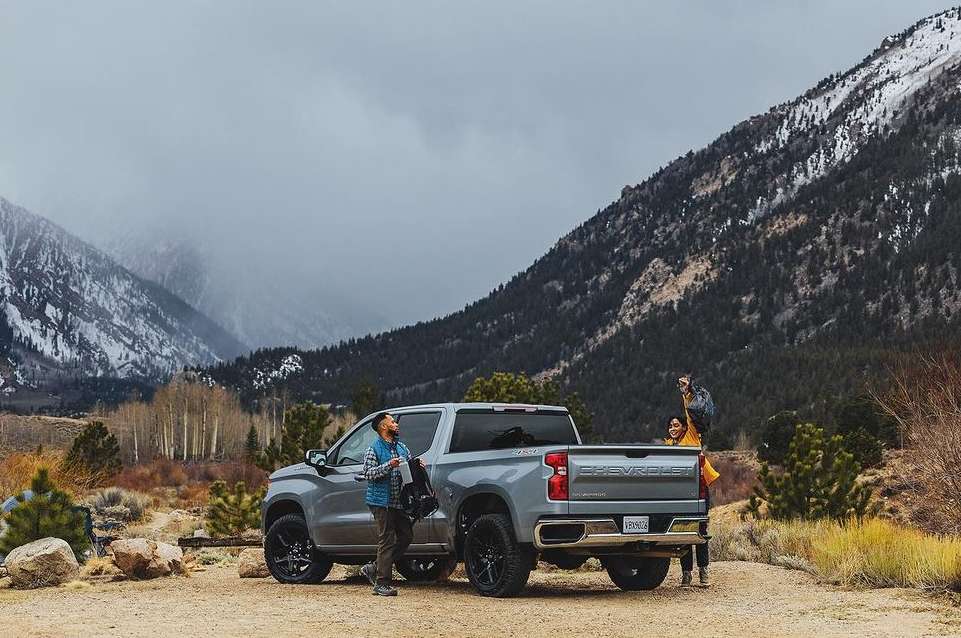 Silver Chevrolet truck parked on a dirt road, with an adult male standing by the driver's side, mountain range in the background.