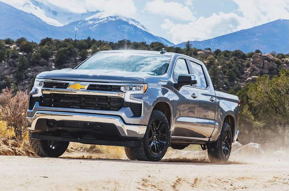 Grey Chevrolet Silverado pickup truck drives on a dirt road with mountains and trees in the background. Automotive industry.