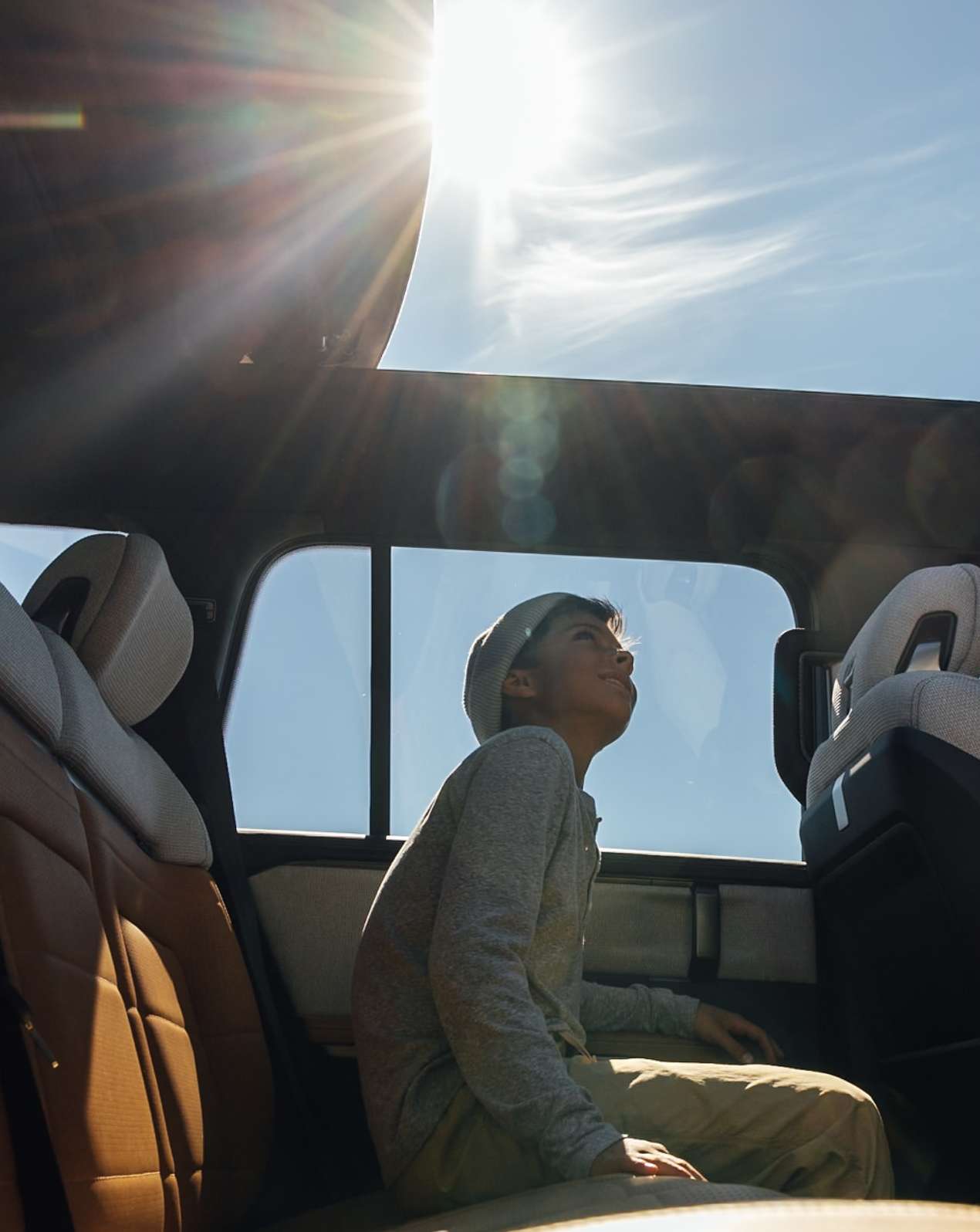 Boy in car looking up at sun through open sunroof, enjoying road trip on a sunny day with blue sky and car interior details.