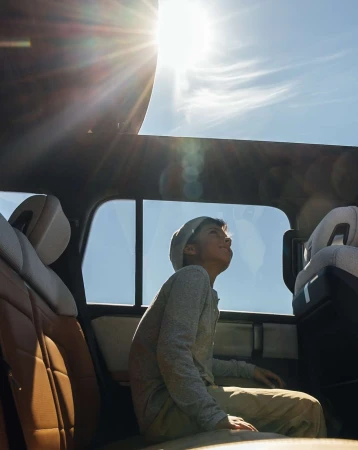 Boy in car looking up at sun through open sunroof, enjoying road trip on a sunny day with blue sky and car interior details.