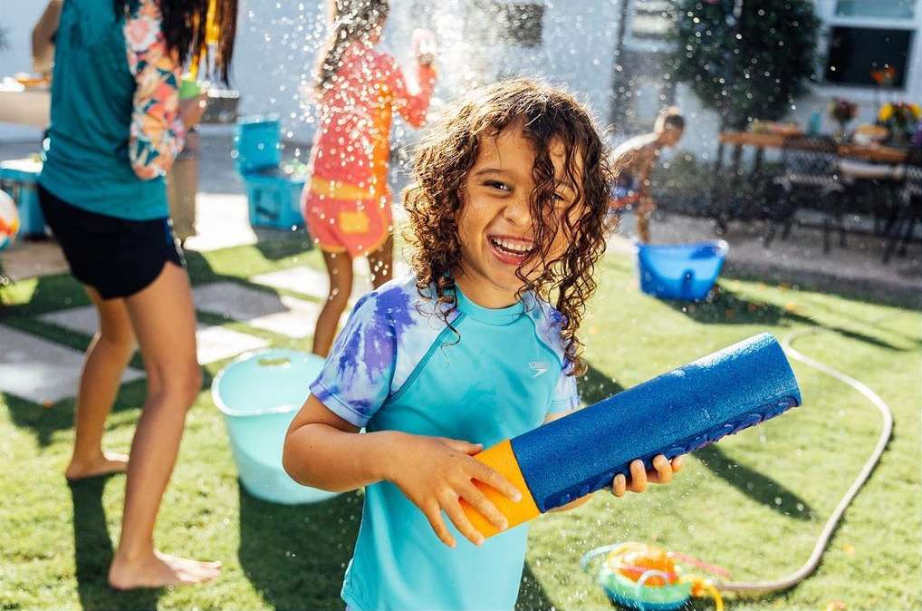 Child smiles, holding a water blaster toy during a water fight in a backyard on a sunny day, surrounded by other kids and water toys.