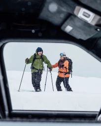 Two skiers with backpacks and ski poles walking through deep snow viewed through a car window frame.