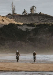 Two riders in helmets driving motorcycles along a sandy beach with steep grassy dunes and sparse trees in the background.