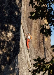 Rock climber in red trousers and white top scaling a steep cliff face with climbing harness and blue helmet