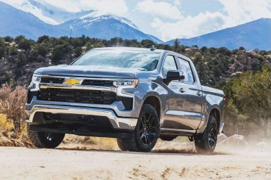 Grey Chevrolet pickup truck driving on a dusty dirt road with snowy mountain peaks in the background.