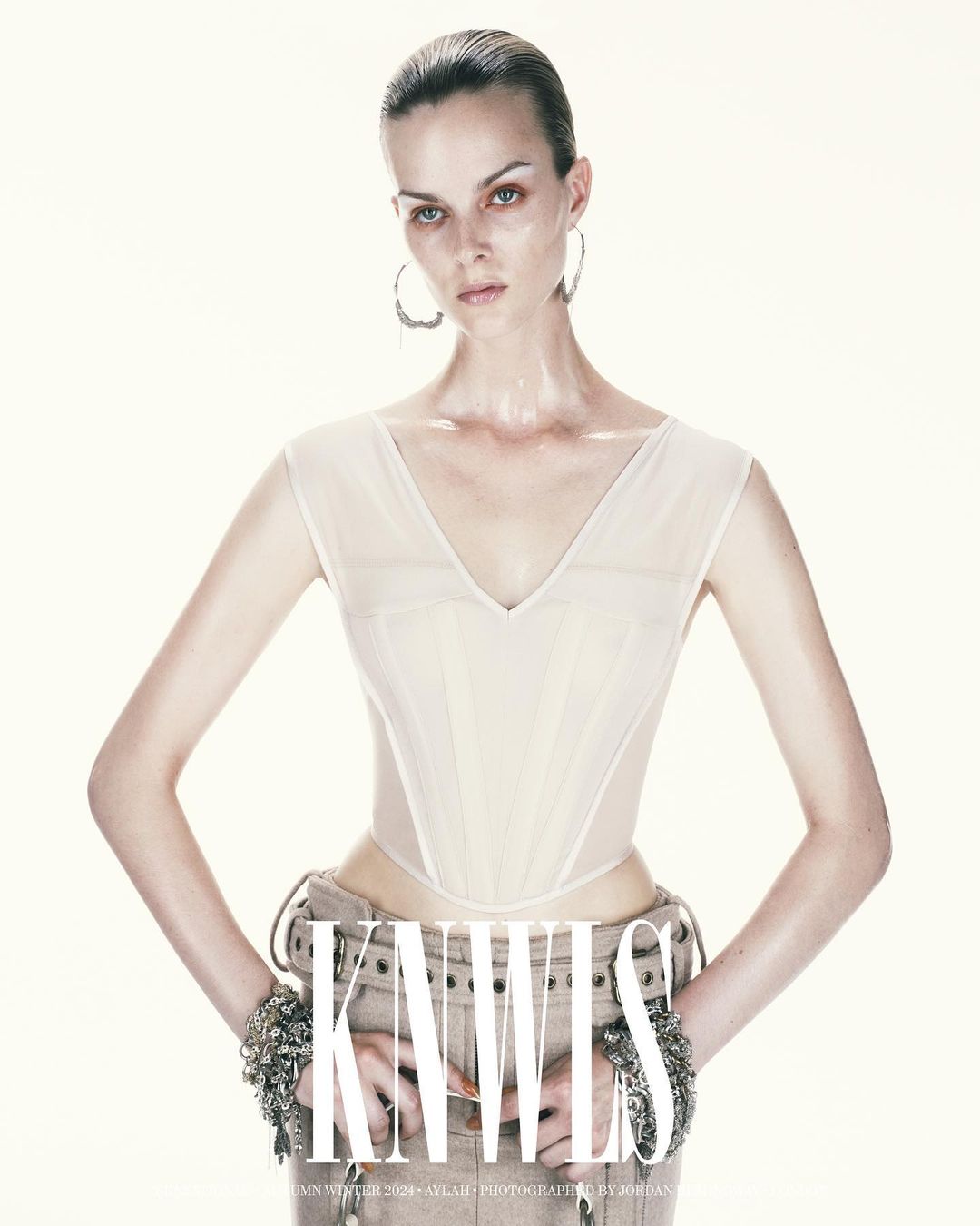A woman poses in an ivory corset top and tan pants with bracelets on a white background, showcasing fashion, studio lighting, and style.