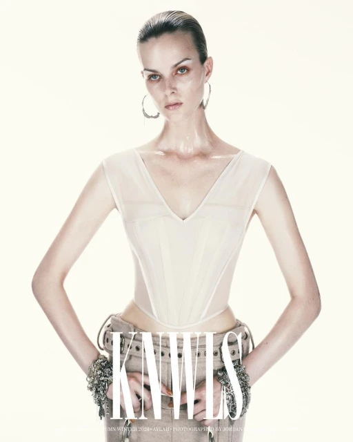 A woman poses in an ivory corset top and tan pants with bracelets on a white background, showcasing fashion, studio lighting, and style.