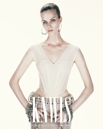 A woman poses in an ivory corset top and tan pants with bracelets on a white background, showcasing fashion, studio lighting, and style.