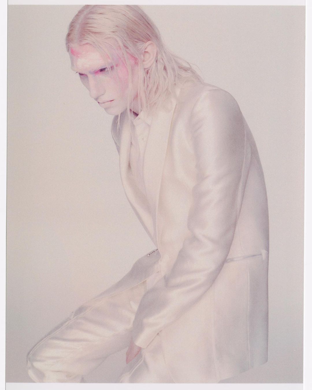 A person with white hair and makeup wears a white suit in a studio shot. The person is leaning forward with their head tilted down.
