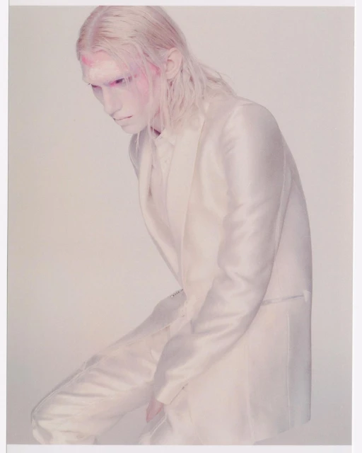 A person with white hair and makeup wears a white suit in a studio shot. The person is leaning forward with their head tilted down.