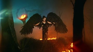 Woman in black dress with large wings stands in a smoky, fiery landscape. Dark tones with orange and blue lighting effects create a dramatic scene.
