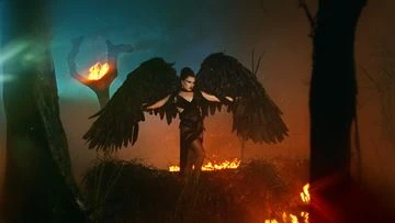 Woman in black dress with large wings stands in a smoky, fiery landscape. Dark tones with orange and blue lighting effects create a dramatic scene.