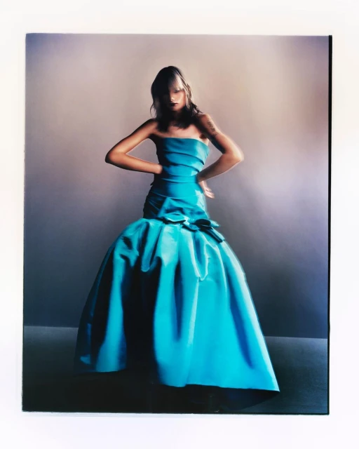 A fair-skinned young adult woman poses indoors wearing a blue strapless formal gown with a full skirt and hands on her hips.