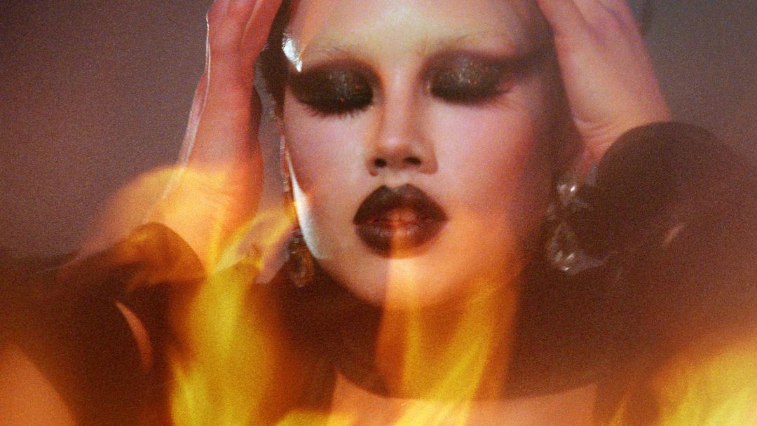 Close-up of a woman with dramatic makeup and her hands raised, bathed in warm, diffused light creating an ethereal visual.