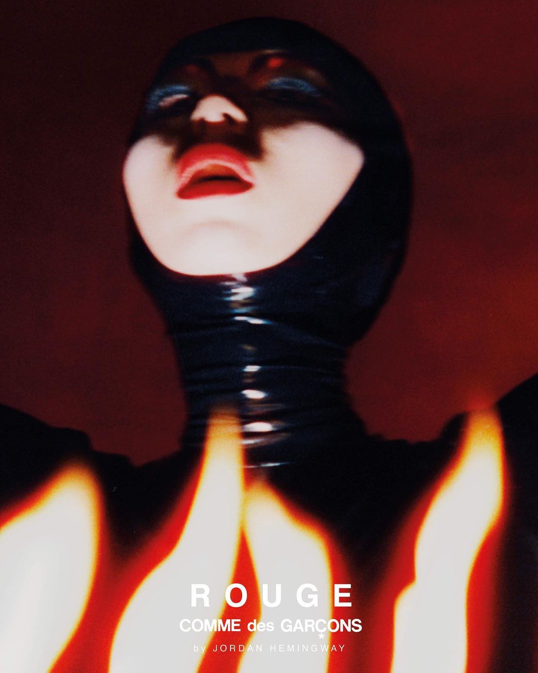 Close-up fashion shot of a person in a black latex hood, with red lipstick, against a dark red backdrop, lit by red and yellow light.