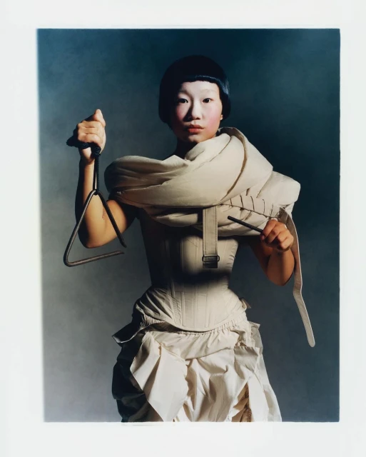 Young woman with pale face paint poses indoors wearing avant-garde white dress, holding musical triangle, and looking at the viewer.