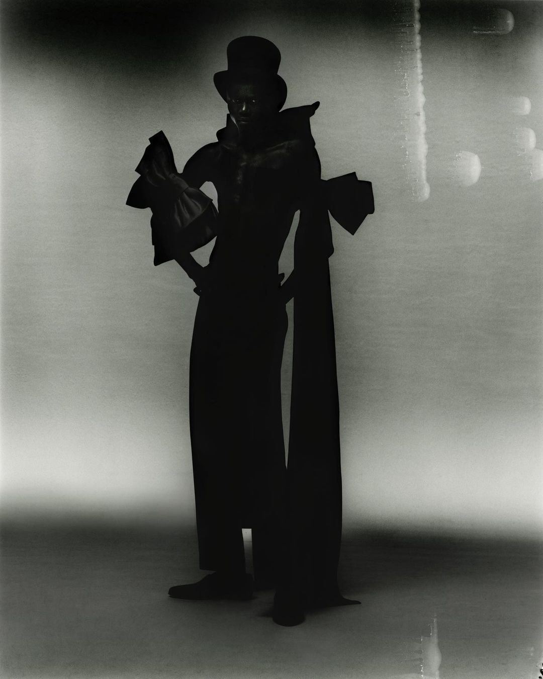 Fashionable figure in dramatic lighting, wearing a top hat and wide-sleeved outfit with long flowing panels, set against a soft background.