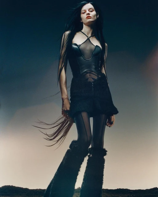 A slender fashion model poses in a black mesh and leather corset-like top, sheer leggings, and fur leg coverings against a studio backdrop.