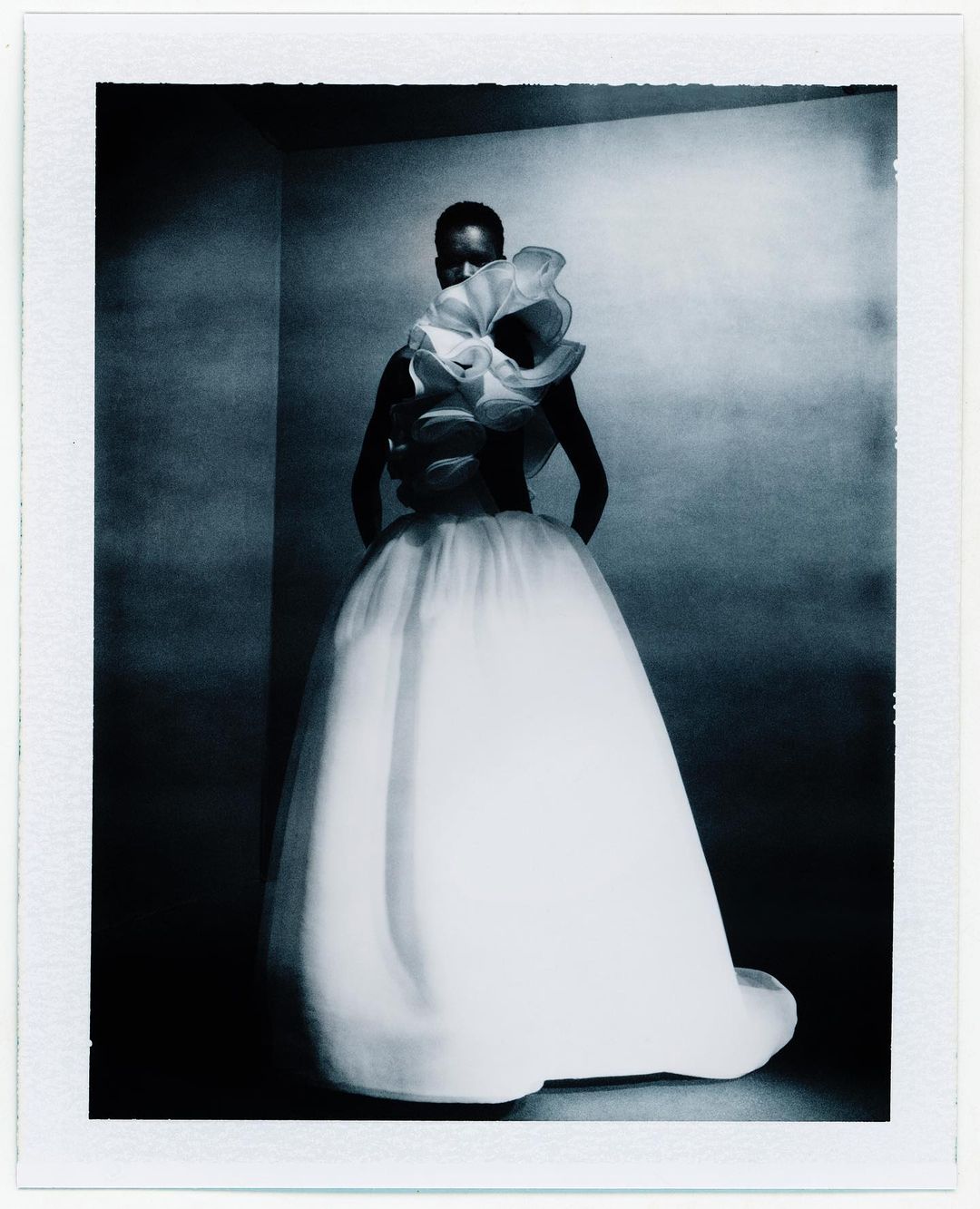 Woman in a white tulle gown with floral ruffles poses in a corner photo studio with dramatic lighting and a polaroid frame effect.