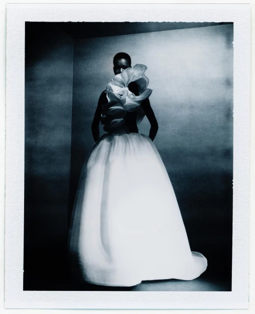 Woman in a white tulle gown with floral ruffles poses in a corner photo studio with dramatic lighting and a polaroid frame effect.
