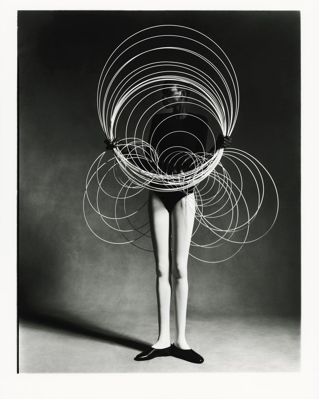 Black and white studio shot of a female model holding a large circular wire sculpture, wearing a bodysuit and flats against a grey backdrop.