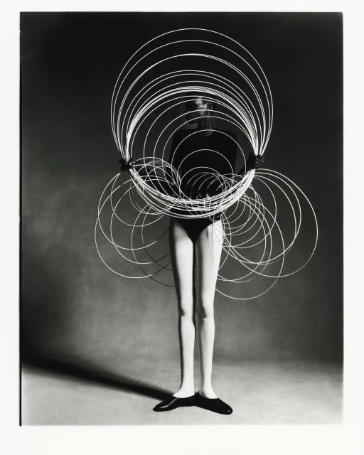 Black and white studio shot of a female model holding a large circular wire sculpture, wearing a bodysuit and flats against a grey backdrop.