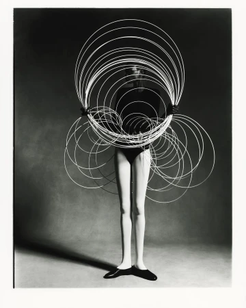 Black and white studio shot of a female model holding a large circular wire sculpture, wearing a bodysuit and flats against a grey backdrop.