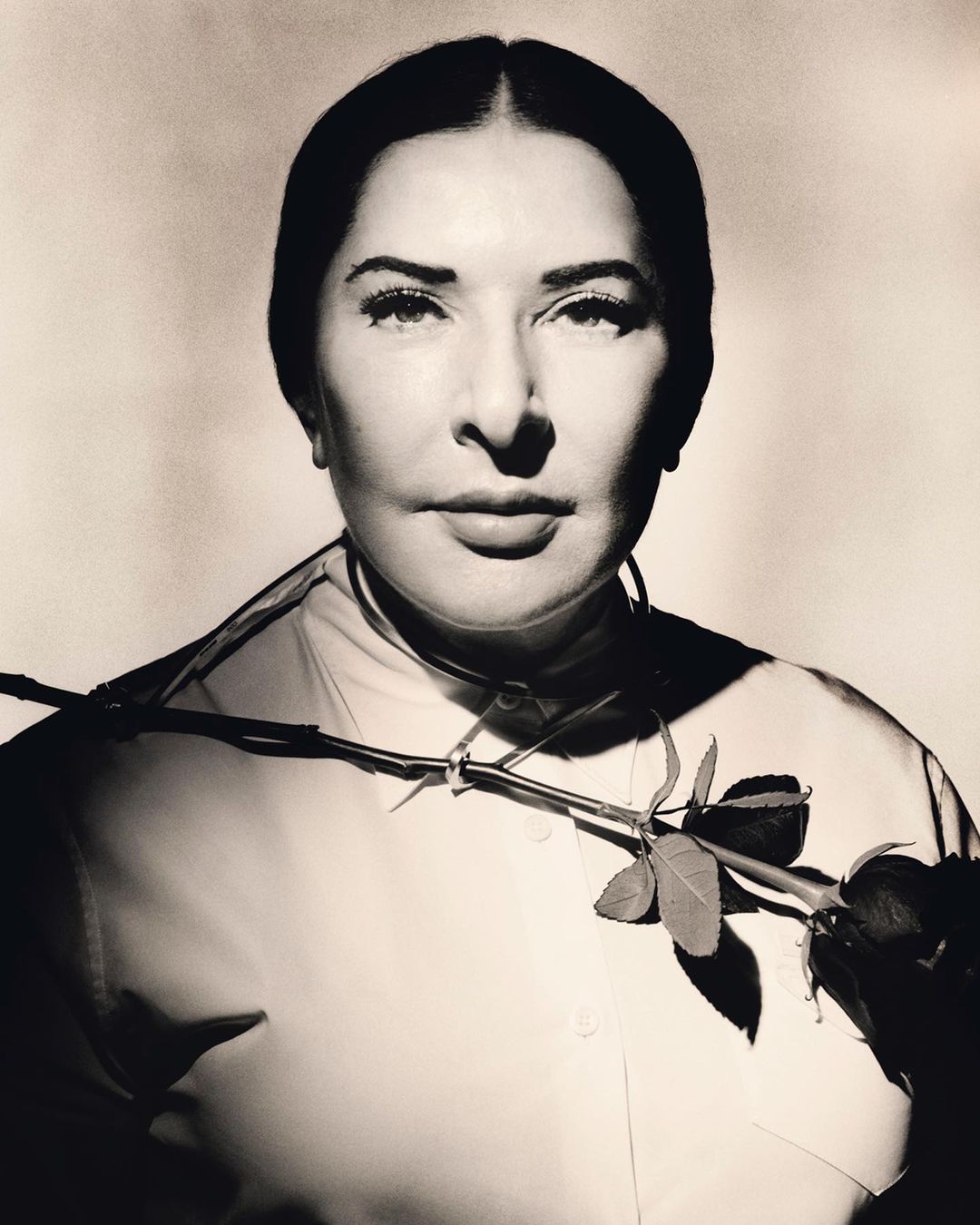 Monochrome portrait of an adult woman with dark hair wearing a collared shirt, adorned with a rose stem across her chest in studio lighting.