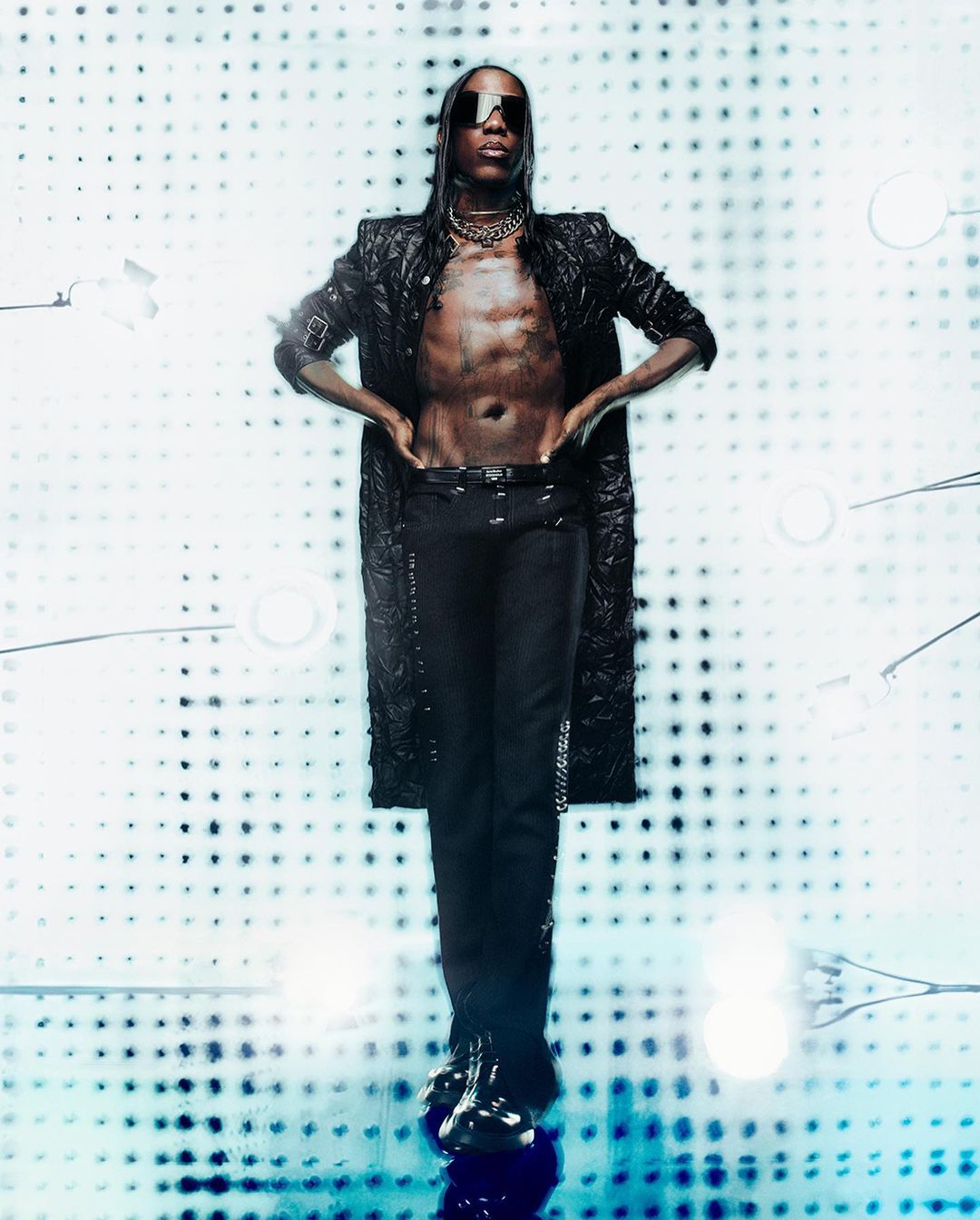 Model wearing dark sunglasses, an unbuttoned black textured coat, and black trousers against a dotted backdrop.