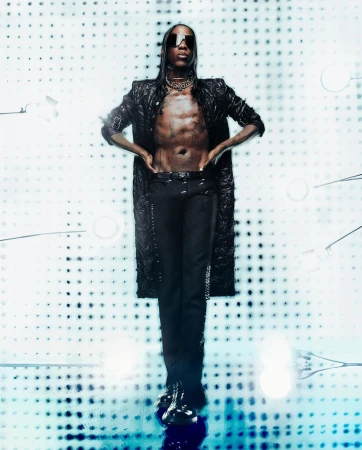 Model wearing dark sunglasses, an unbuttoned black textured coat, and black trousers against a dotted backdrop.