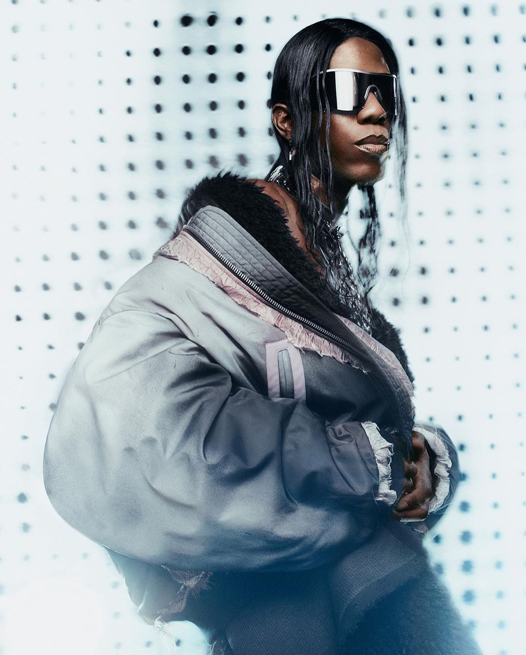 Person with long dark hair poses in a stylish jacket and futuristic sunglasses against a dotted white background in a portrait shot.