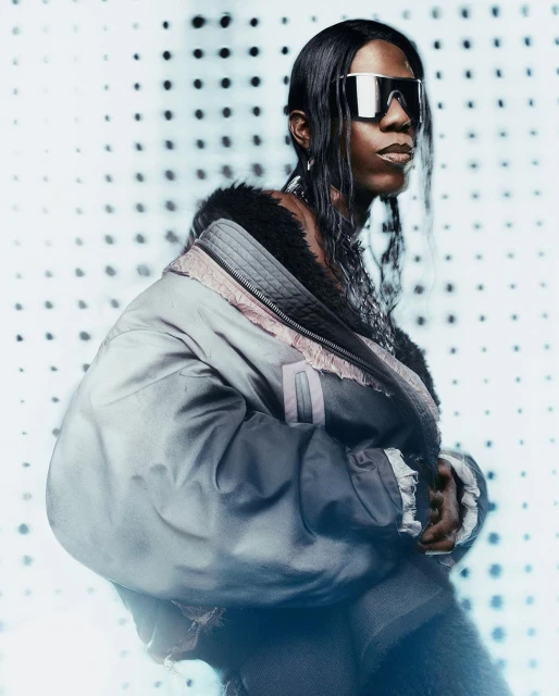 Person with long dark hair poses in a stylish jacket and futuristic sunglasses against a dotted white background in a portrait shot.