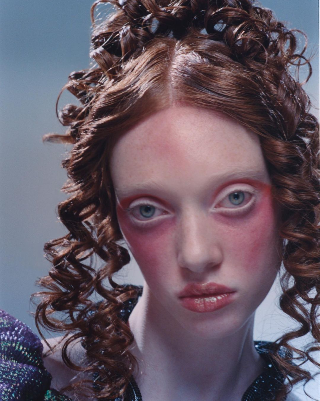 Close-up of a young adult female fashion model with elaborate curly brown hair, striking face makeup, light eyes, and ornate dress.