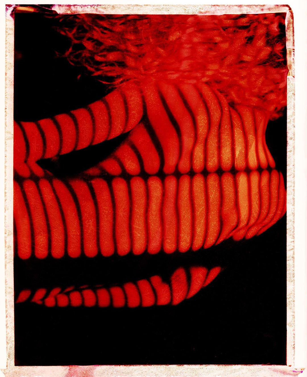 Abstract portrait of a reclining nude female figure with wavy red hair, photographed with linear orange and black striped lighting.