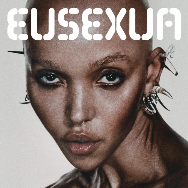 Close-up portrait of a bald woman with clear spike earrings, a nose ring, and large text reading 'EUSEXUA' across the top.