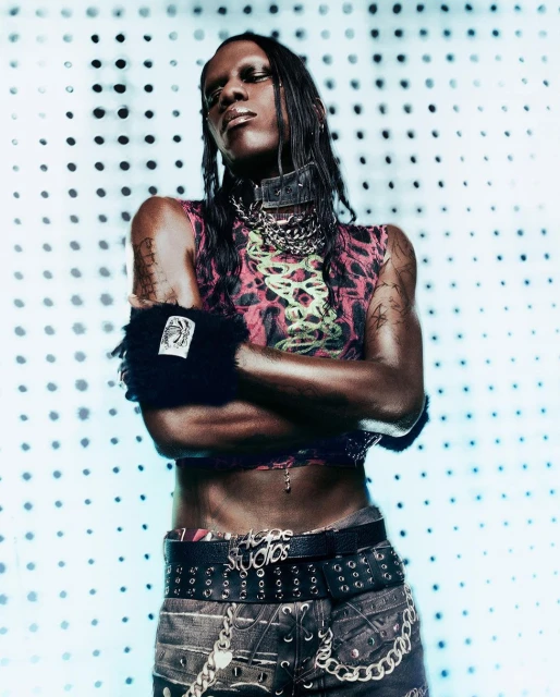 Adult in avant-garde fashion poses in a pink top, black leather collar, jeans, and black cuff on a blue-and-white polka dot background.