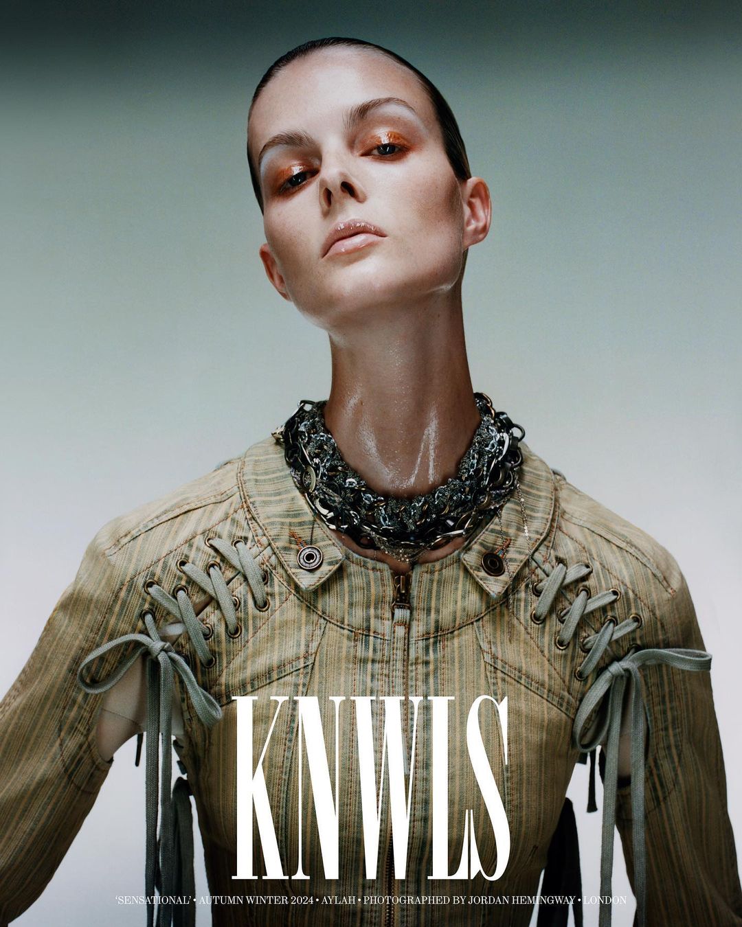 Fashion portrait of a woman with a shaved head wearing a jacket and chain necklace in a photo studio with diffused lighting.