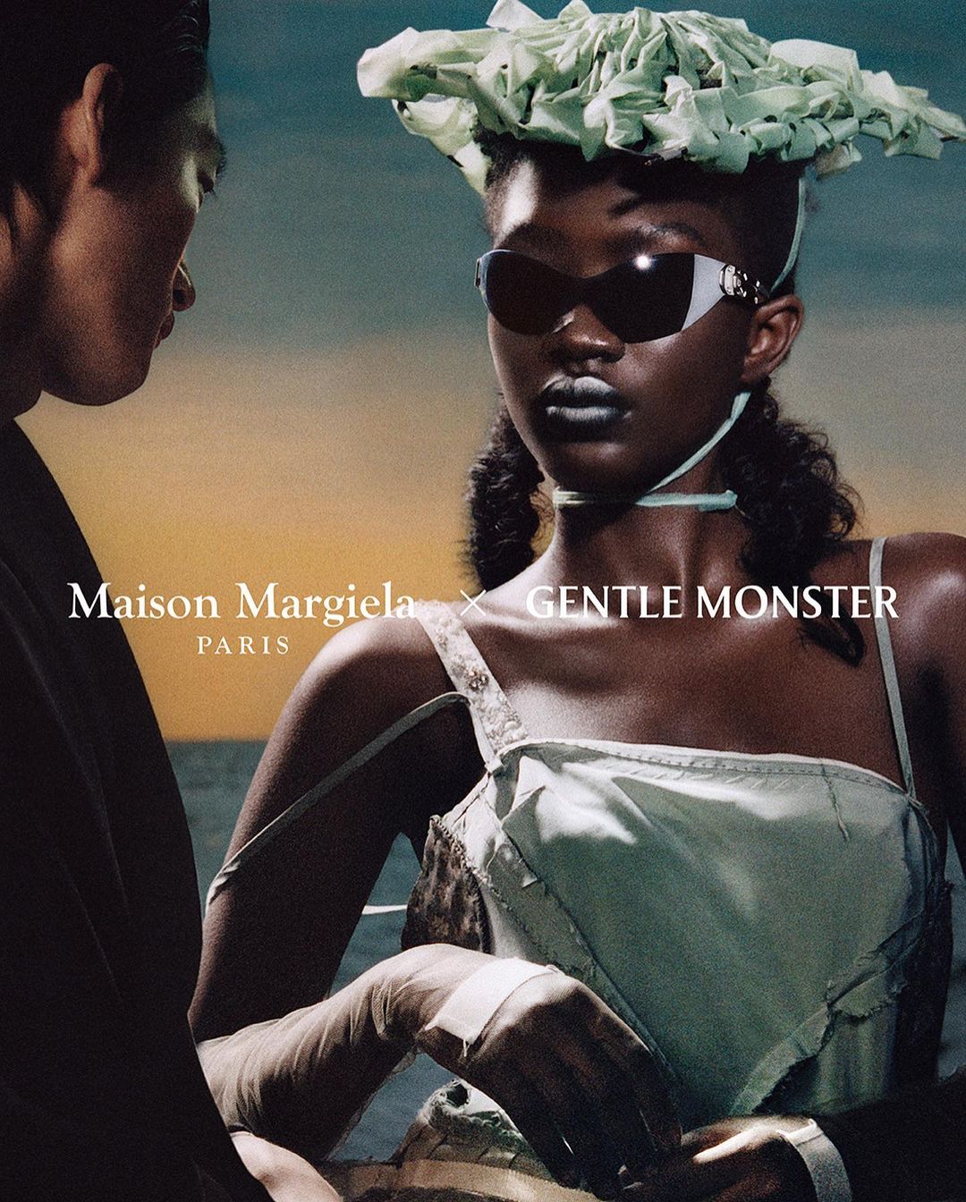 Eye-level shot of two models. A woman wears light-green clothing, sunglasses, and a sculptural hat. Text reads Maison Margiela x Gentle Monster.
