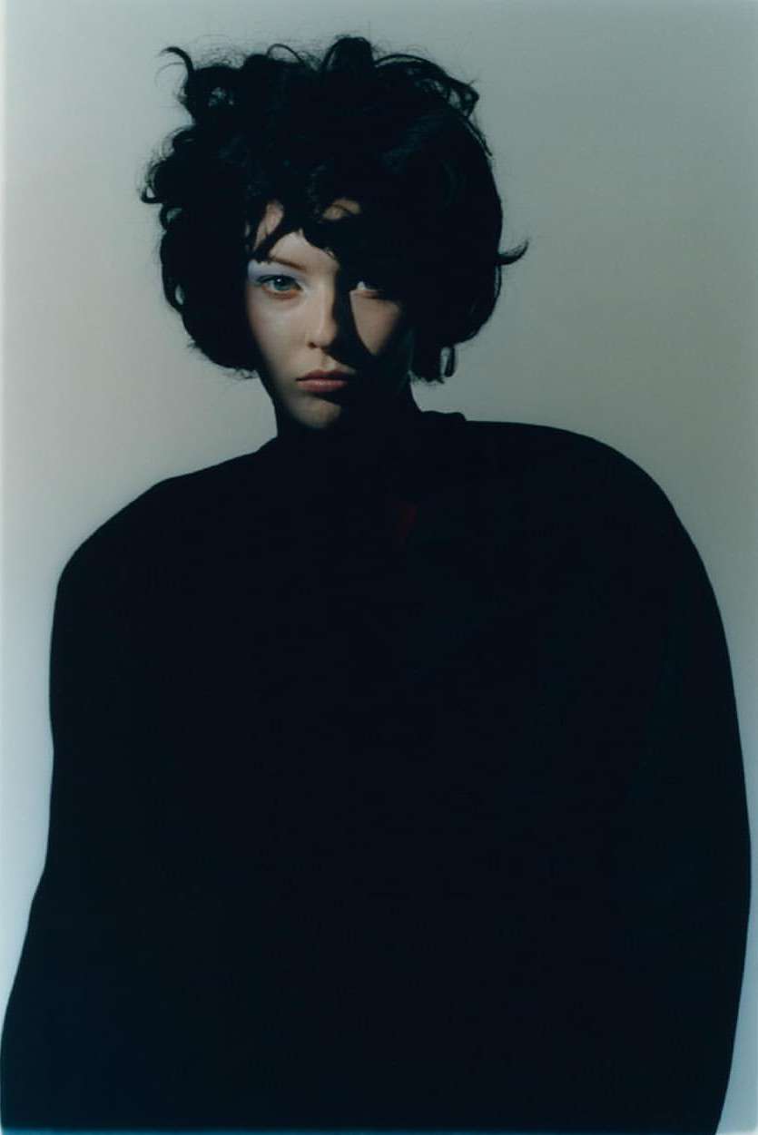 Portrait of a woman with dark, curly hair and pale skin, wearing a black sweater, posing in a photo studio with soft lighting.