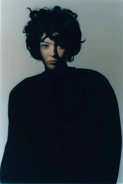 Portrait of a woman with dark, curly hair and pale skin, wearing a black sweater, posing in a photo studio with soft lighting.