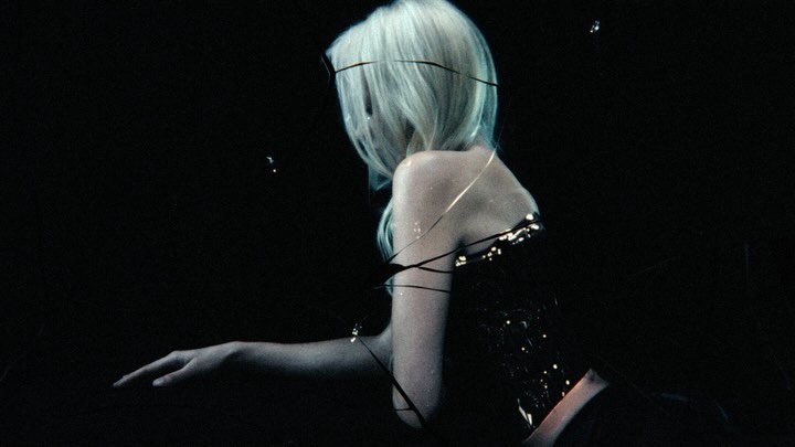 Adult woman with long blonde hair sits in the dark wearing a shiny dark bustier, arm outstretched, dramatic lighting and mysterious feel.