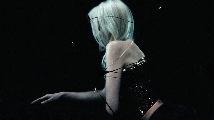 Adult woman with long blonde hair sits in the dark wearing a shiny dark bustier, arm outstretched, dramatic lighting and mysterious feel.
