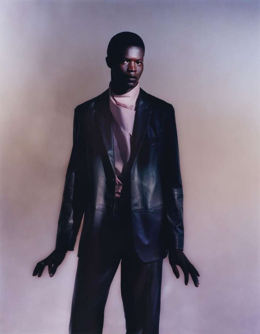 Portrait of a Black male model in a leather blazer and lavender scarf posing in front of a neutral colored backdrop in a studio.
