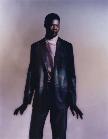 Portrait of a Black male model in a leather blazer and lavender scarf posing in front of a neutral colored backdrop in a studio.